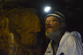A man in a white jumpsuit and a light on his helmet inspects cave crevices for bats. 