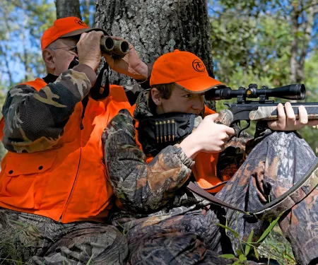 Man looking through binoculars while young boy looks through scope of the rifle out hunting.  Both wearing hunter orange vest and hat.