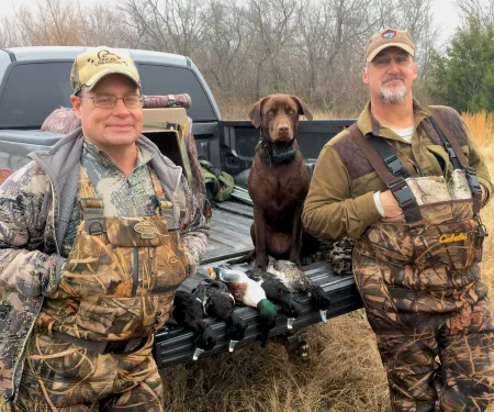 Two men stand next to a truck with a dog and five ducks on a tailgate. 