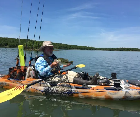 A person holds a fish while sitting in a kayak with fishing gear on the water. 