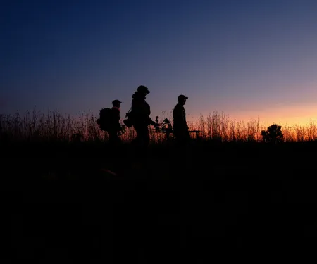 A group of archery hunters are walking through a field at sunrise.
