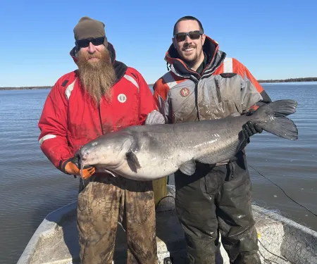 Two biologists in coats hold a big catfish while standing on a boat. 
