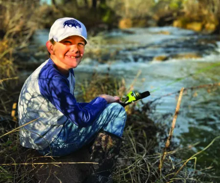A young man sits at the edge of a waterway with a fishing pole in hand. 