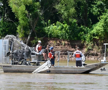 Three people navigate a river on an airboat with nets hanging over the sides of the boat. 