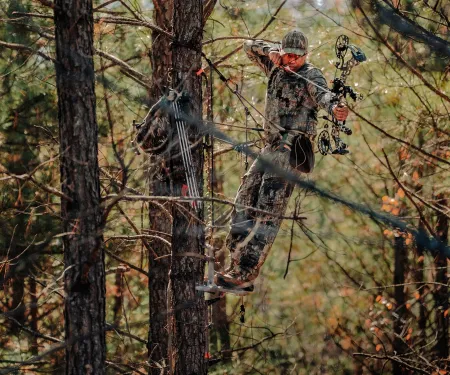 A bowhunter is in a sling tree stand while aiming his bow downward.