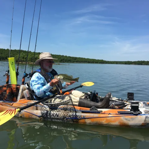 A person holds a fish while sitting in a kayak with fishing gear on the water. 