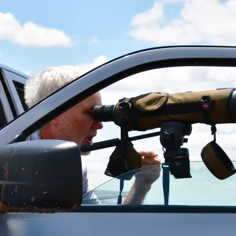 A man looks through a spotting scope attached to an open truck door. 