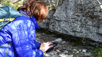 A woman in a blue coat leans over a GPS unit to document a plant growing on a rock ledge.