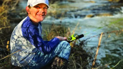 A young man sits at the edge of a waterway with a fishing pole in hand. 