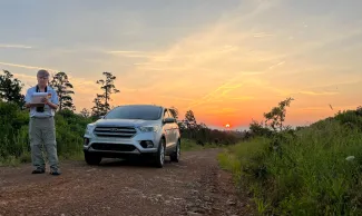 A biologist stands in front of a vehicle at daybreak with a data sheet in hand and binoculars and stop watch around his neck. 