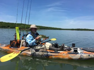 A person holds a fish while sitting in a kayak with fishing gear on the water. 