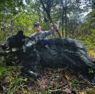 A man with a bow in hand in front of a harvested black bear. 