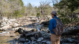 A man with a backpack stands near a rocky stream. 