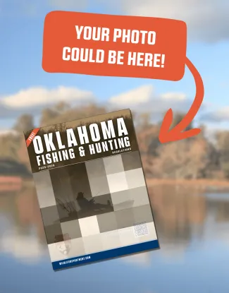 A promotional graphic showing a blurred outdoor lake scene in the background. At the top, an orange speech-bubble banner with an arrow reads “YOUR PHOTO COULD BE HERE!” Below it, an angled mock-up of the Oklahoma Fishing & Hunting Regulations 2025–2026 booklet is displayed, featuring a placeholder grid image of a person fishing from a kayak.