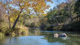 A photograph of Blue River in Oklahoma. The water is reflective and the trees are turning to fall colors.