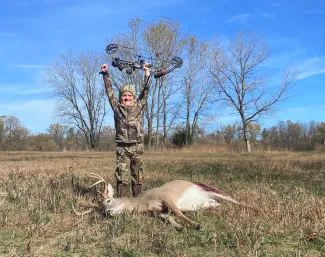 A youth bow hunter stands triumphantly holding their bow above their head behind their harvested deer.