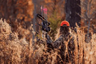 A female bowhunter walks through tall grass while wearing a hunter orange hat.